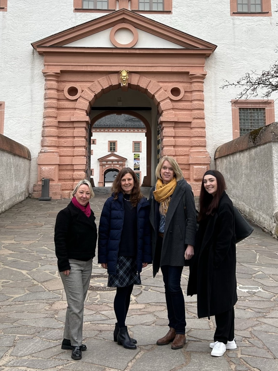 Vier lächelde Frauen vor einen historischen Torbogen eines historischen Gebäudes.